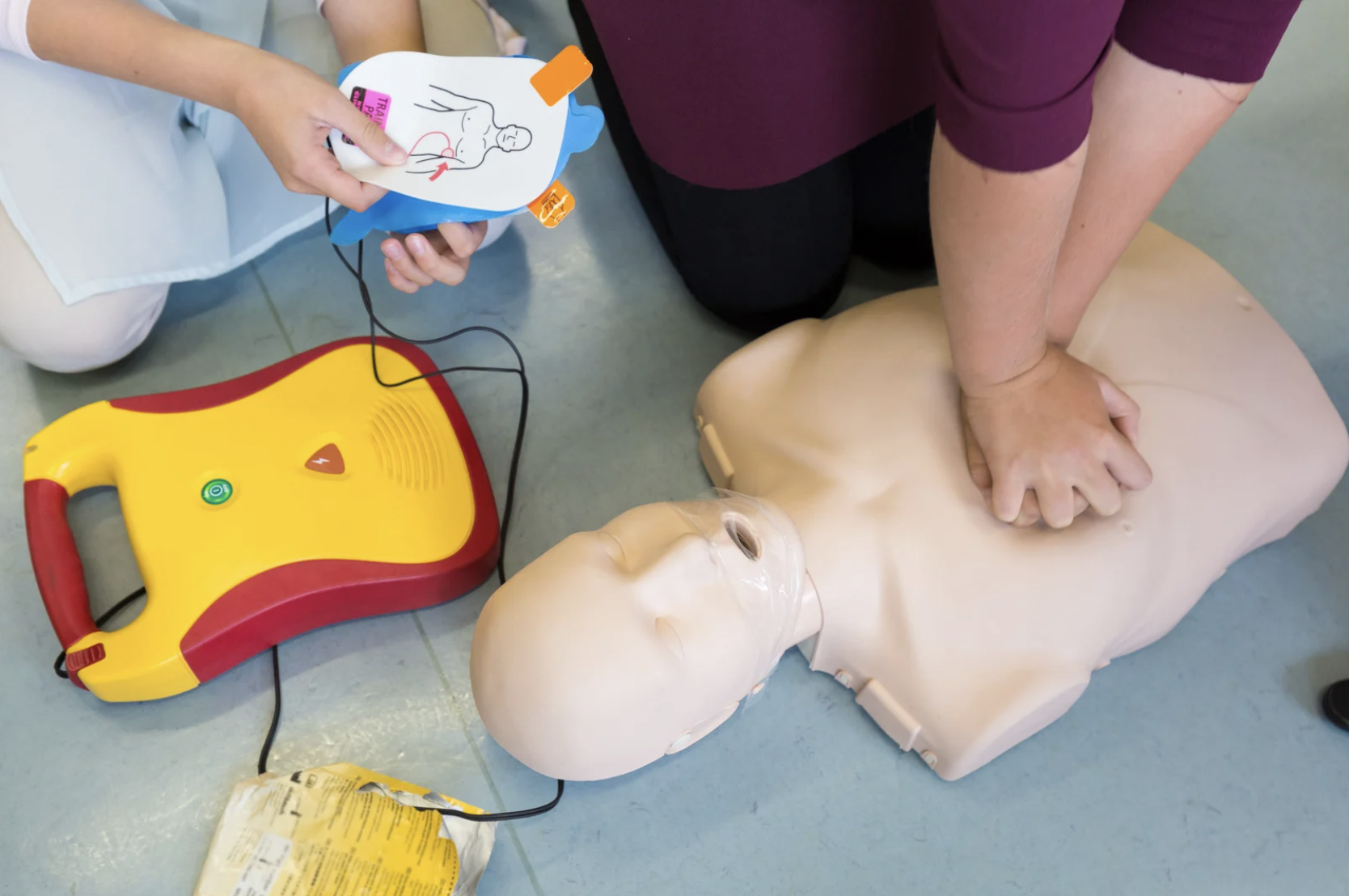 Hands practising CPR on a mannequin beside an AED trainer