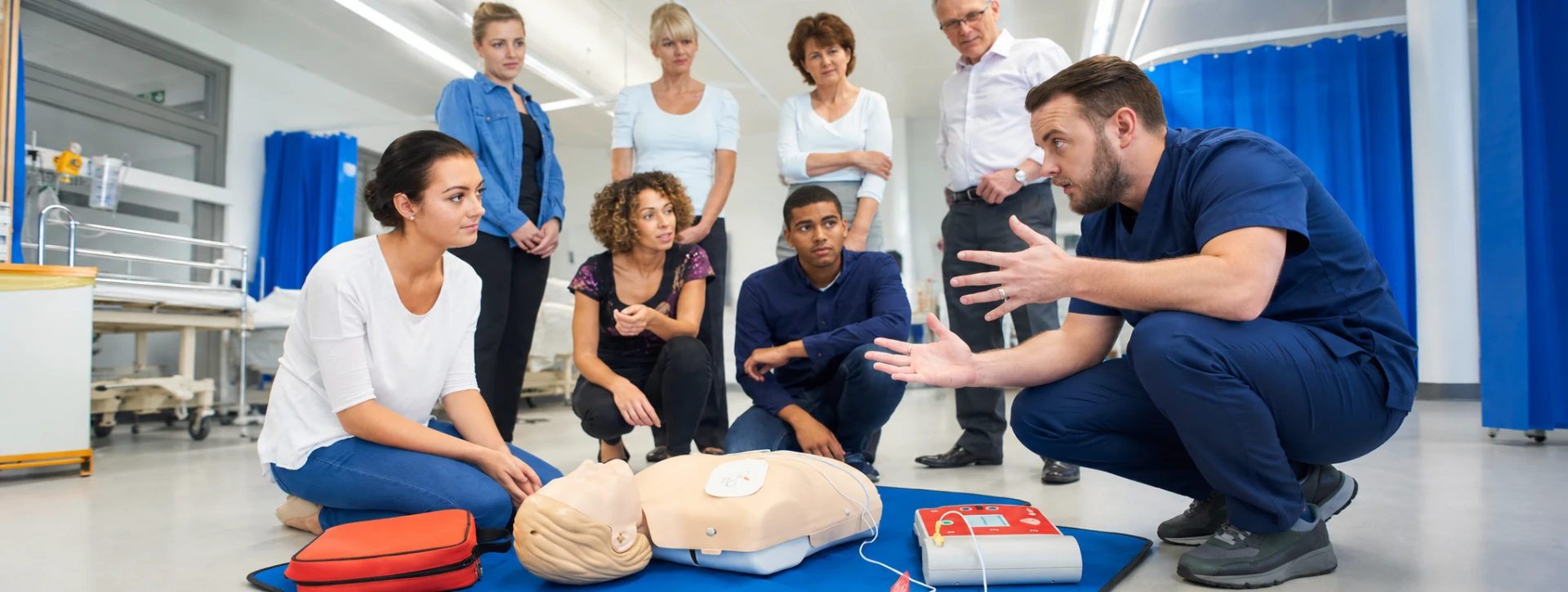 An instructor explaining CPR and defibrillator use to a group gathered around a training mannequin